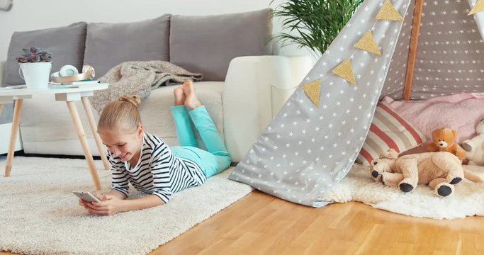 Child girl using cell phone lying on the carpet indoors near sofa and smiling at camera
