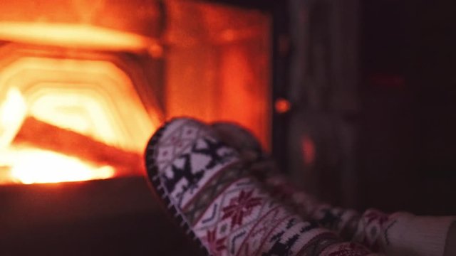 Feet in woollen socks by Burning Christmas Fireplace. 4K STABILIZED shot. Woman relaxes by warm fire with a cup of hot drink and warming up her feet. Close up. Winter and Christmas holidays concept.