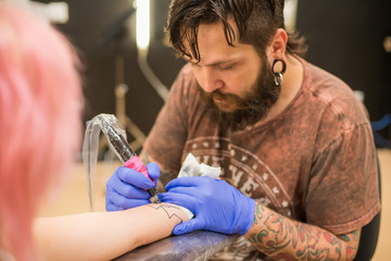 A bearded tattoo artist , in a brown t-shirt fills the hand tattoo the girl with the pink hair and tattoos and piercings all over her body