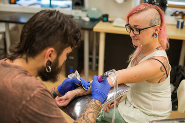 A bearded tattoo artist , in a brown t-shirt fills the hand tattoo the girl with the pink hair and tattoos and piercings all over her body