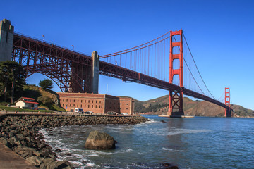 Fototapeta premium Golden Gate bridge from the San Francisco side. View of Fort Mason on the left and rocks in the foreground. A blue sky is in the background. It is a horizontal image.