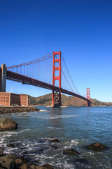 Naklejka premium Golden Gate bridge from the San Francisco side. View of Fort Mason on the left and rocks in the foreground. A blue sky is in the background. It is a vertical image.