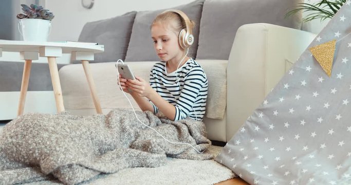 Cheerful child girl listening music in headphones and dancing sitting on the carpet