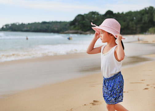 Cute little asian girl wear straw hat walking on the beach with looking out to the sea.