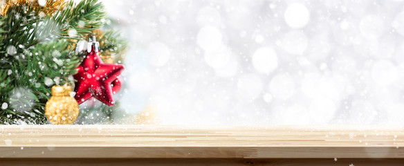 Fototapeta na wymiar Wood table top Christmas holiday banner background with blurred decorated pine tree, white bokeh and snow falling in background