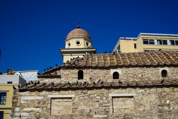 Old classic little church in earth tone natural stone with pigeons on terracotta roof tile with clear blue sky and modern building background