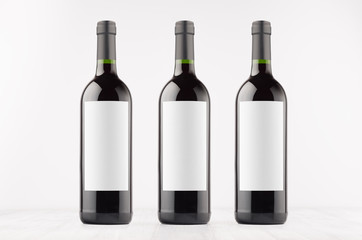 Three dark wine bottles with blank white labels on white wooden board, mock up. Template for advertising, design, branding identity.