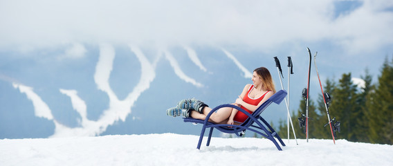 Panoramic view. Cheerful female skier wearing red bodice, lying on a blue deck chair near skis and poles at winter ski resort on the top of the hill. Mountains, ski slopes and forest on the background