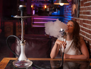 Beautiful girl in a white blouse smokes a hookah in the interior of the bar