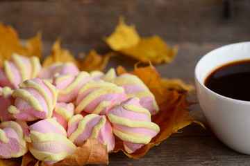 Light pink and yellow marshmallow zephyr on yellow autumn leaves, coffee cup on an old wooden background. Nice autumn coffee break concept for women. Closeup