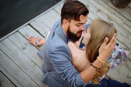 A picture of a young romantic couple. Engagement. Man kissing girl. Thei sitting on desk platform on water of lake.