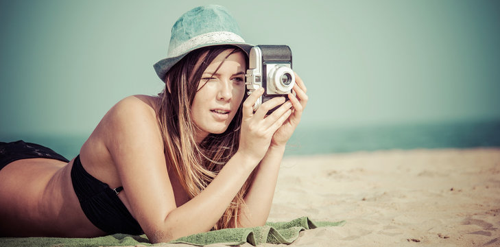 sexy woman lying on beach taking photo with vintage camera. single female tourist on idyllic beach vacation. retro style color filtered wide close up image.