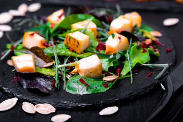 Warm pumpkin salad with mixed leaf of arugula, chard, in on black slate plate on black background. Grilled pumpkin. Close up.