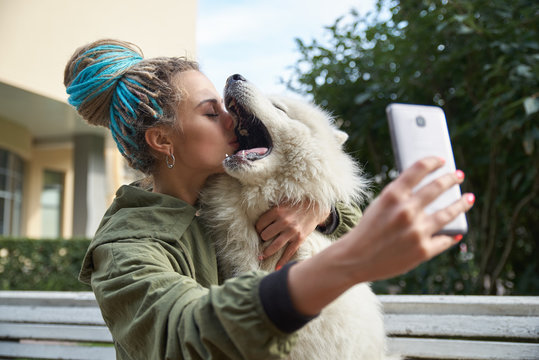 young girl in green coat and multicolored dreadlocks makes a selfie on your smartphone posing with white Samoyed dog in the autumn Park