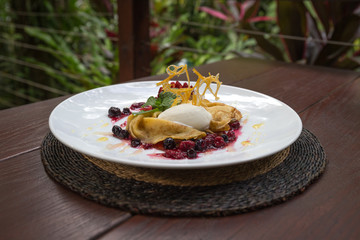 Crepes and ice cream on a white plate with a rainforest garden in the background. The dish contains a nice decoration made of caramelised oranges.