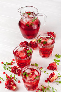 Pomegranate cocktail with ice and mint in beautiful glasses and jug, fresh ripe pomegranate on white wooden background. Sweet red juice. Close up photography. Selective focus