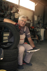 Senior man in workshop. Man sitting on tires and holding his notes. Looking at camera.