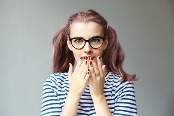 Trendy looking 18 year old girl with pinkish ponytails wearing cat eye glasses and striped t-shirt covering mouth with both hands, having astonished look, shocked with unbelievable information