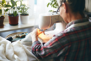 Back view of unrecognizable elderly needlewoman in plaid shirt sewing kitchen curtains using...