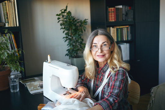 Candid shot of beautiful smiling 60 year old female tailor wearing rectangular glasses and plaid shirt enjoying work on new sewing machine, feeling happy while bringing to life ideas and creativity