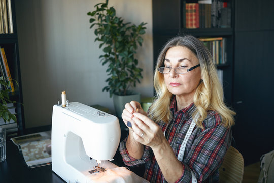 Picture of serious confident blonde retired woman dressmaker in stylish eyewear making custom clothing for women, sitting at her workplace, busy stitching dress using modern white sewing machine