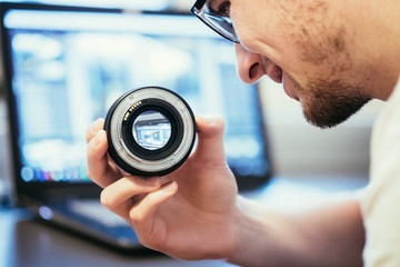 Junger Mann hält Objektiv in der Hand, Laptop im Hintergrund