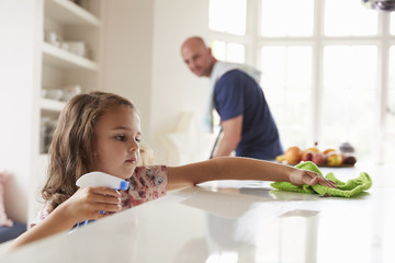 Girl cleaning kitchen worktop, father sweeping floor in the background