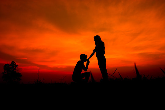 silhouette of a romantic couple on beauty sunset. A man kneels kissing a girlfriend's hand.
