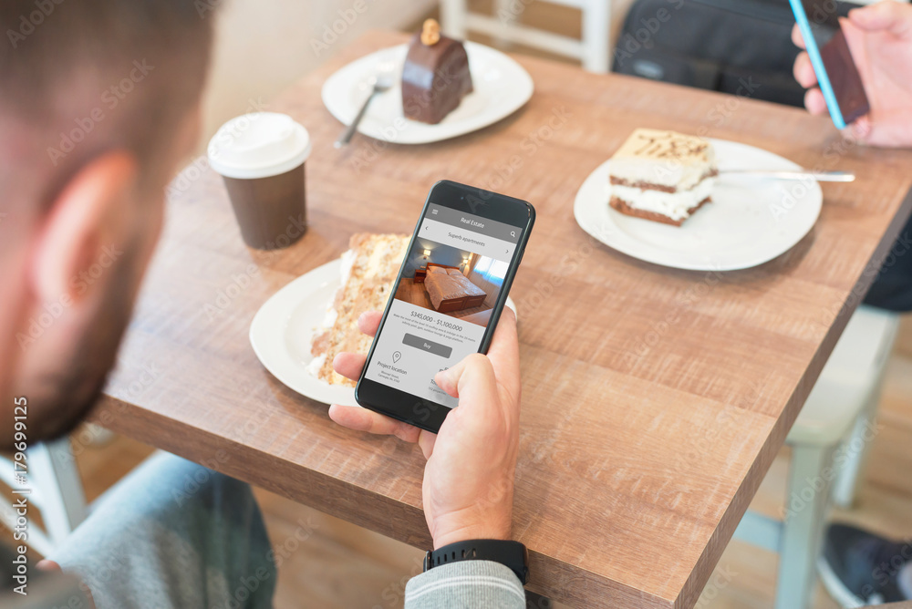Poster Man use real estate app on mobile phone. Coffee and cake on table in background.