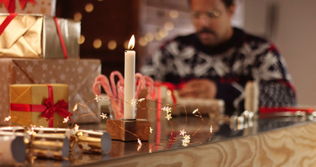 Handsome dark skinned man in black white and red fairisle sweather wrapping Christmas present among decorations and lights