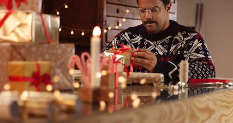 Handsome dark skinned man in black white and red fairisle sweather wrapping Christmas present among decorations and lights