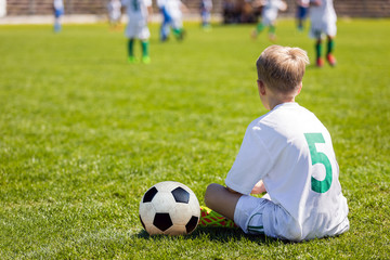 Young Boy as a Soccer Player. Kid Sitting on Football Pitch. Soccer Stadium in the Background. Youth Soccer Match. Football School Tournament Competition