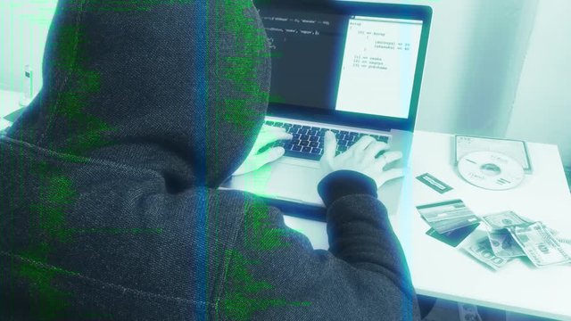 Anonymous Hacker Attacks In Cyberspace, Zoom In. Hooded person typing in a laptop next to stolen itens, simulating a hacker crime scene