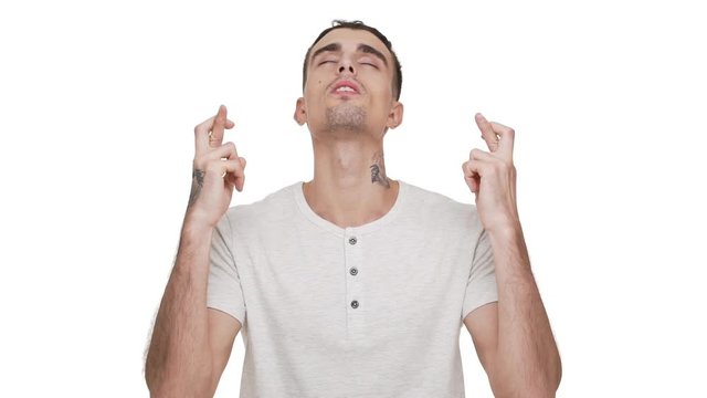 slow motion portrait of praying young man wearing casual shirt wishing something good looking up with closed eyes keeps fingers crossed begging god please over white background closeup. Concept of