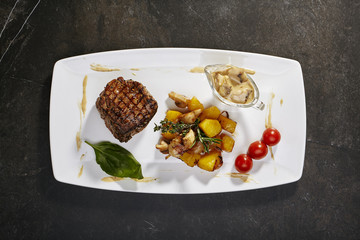 Steak filet mignon with garnish of fried potatoes with porcini mushrooms and creamy mushroom sauce with greens on black background