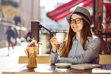 Stylish cheerful girl wears blue shirt, hat and eyeglasses having cup of coffee in street cafe, outdoors sunny autumn time
