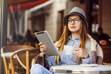 Fototapeta premium Stylish cheerful girl wears blue shirt, hat and eyeglasses, plaid, having cup of coffee using tablet pc in street cafe, outdoors sunny autumn time