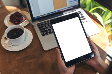 Mockup image of hands holding black tablet pc with blank white screen , laptop , coffee cup and cake on wooden table in cafe