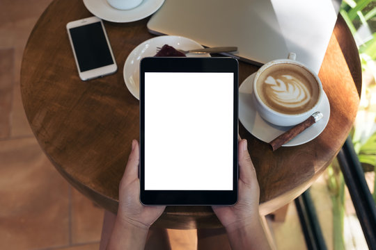 Top view mockup image of hands holding black tablet pc with blank white screen , smart phone , laptop , coffee cup and cake on wooden table in cafe