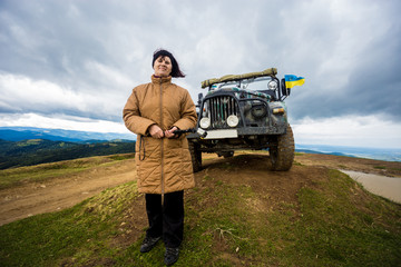 Happy senior tourist woman travel by jeep in mountain forest in autumn with dramatic sky on background