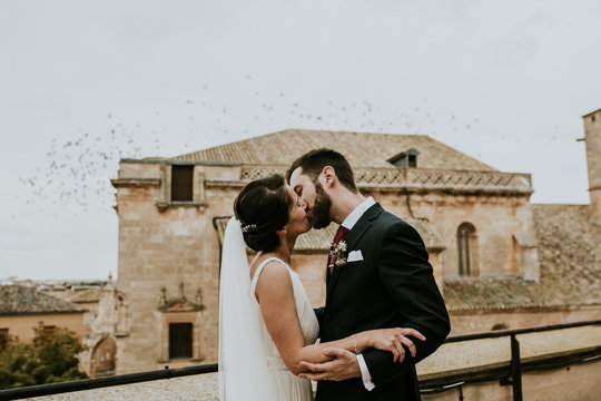 .Young and beautiful couple in love taking pictures on their wedding day kissing on a roof and pigeons flying in the background. Celebrating their marriage. Lifestyle photography.
