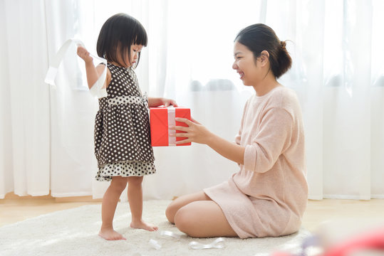 Asian Little Girl And Her Mother Unwrapping A Red Gift Box Together.