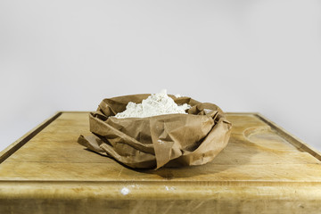 Brown paper sack of white flour in the center of rough wooden rectangular cutting board from side