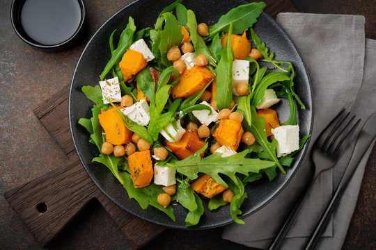 Vegetarian salad with arugula, baked pumpkin, herbs and seasonings, feta cheese, chickpeas and olive oil in a black ceramic plate on a dark concrete or stone background. Selective focus.