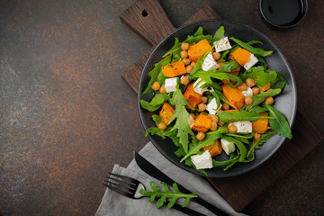 Vegetarian salad with arugula, baked pumpkin, herbs and seasonings, feta cheese, chickpeas and olive oil in a black ceramic plate on a dark concrete or stone background. Selective focus.