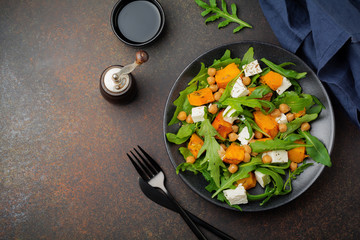 Vegetarian salad with arugula, baked pumpkin, herbs and seasonings, feta cheese, chickpeas and olive oil in a black ceramic plate on a dark concrete or stone background. Selective focus.