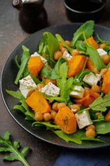 Vegetarian salad with arugula, baked pumpkin, herbs and seasonings, feta cheese, chickpeas and olive oil in a black ceramic plate on a dark concrete or stone background. Selective focus. Top view.