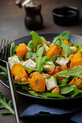 Vegetarian salad with arugula, baked pumpkin, herbs and seasonings, feta cheese, chickpeas and olive oil in a black ceramic plate on a dark concrete or stone background. Selective focus. Top view.