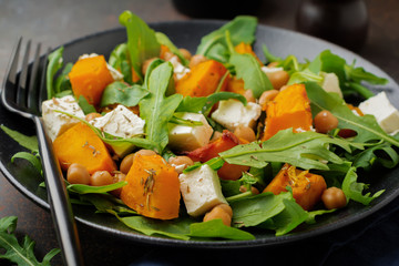 Vegetarian salad with arugula, baked pumpkin, herbs and seasonings, feta cheese, chickpeas and olive oil in a black ceramic plate on a dark concrete or stone background. Selective focus. Top view.