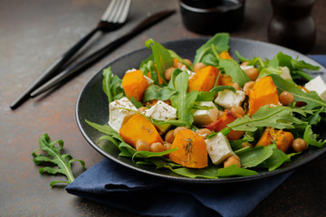 Vegetarian salad with arugula, baked pumpkin, herbs and seasonings, feta cheese, chickpeas and olive oil in a black ceramic plate on a dark concrete or stone background. Selective focus. Top view.